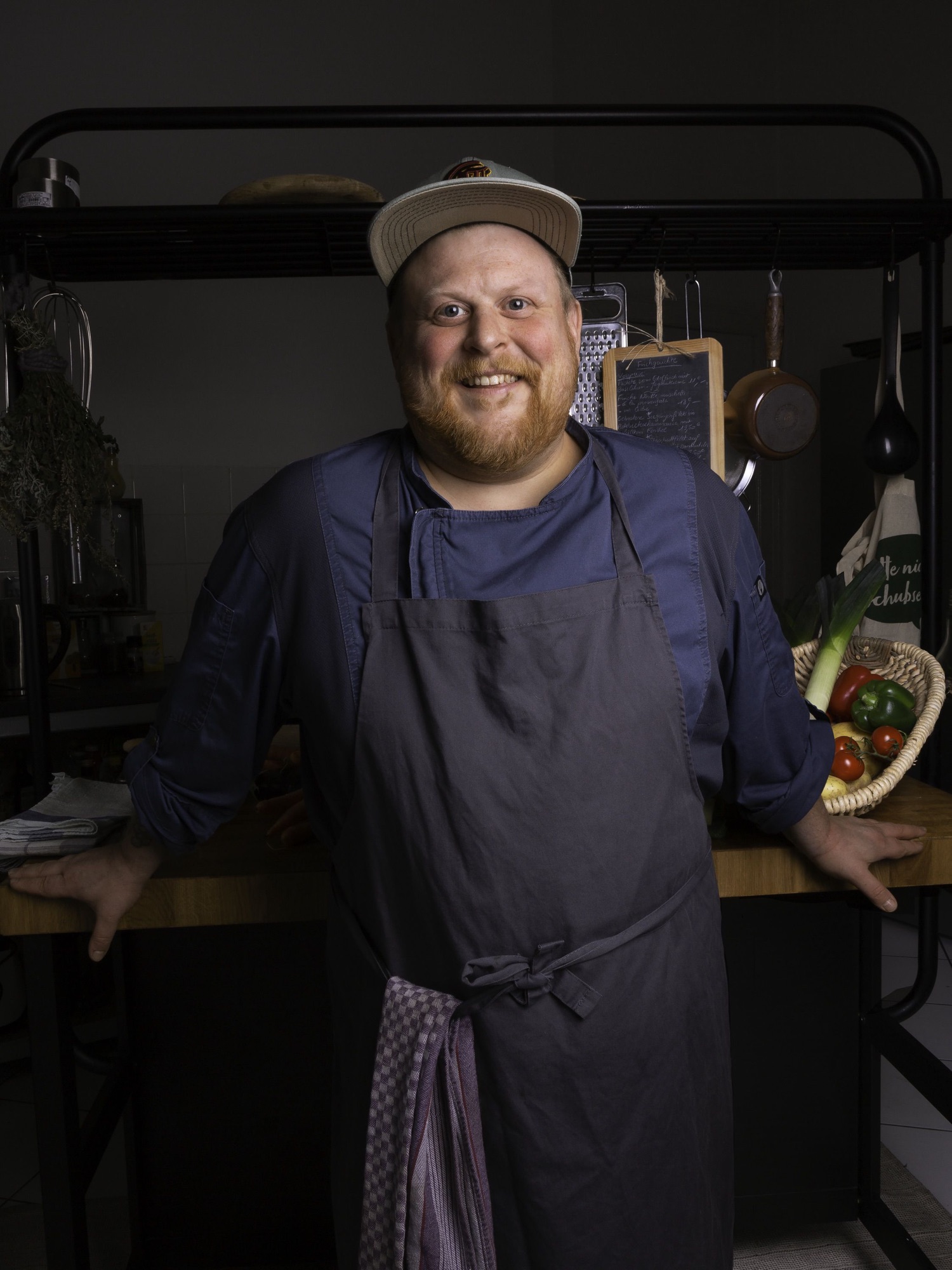 Timo Franke mit Mütze und Schürze in seiner Küche, hinter ihm Kreidetafel, Reibe und ein Gemüsekorb